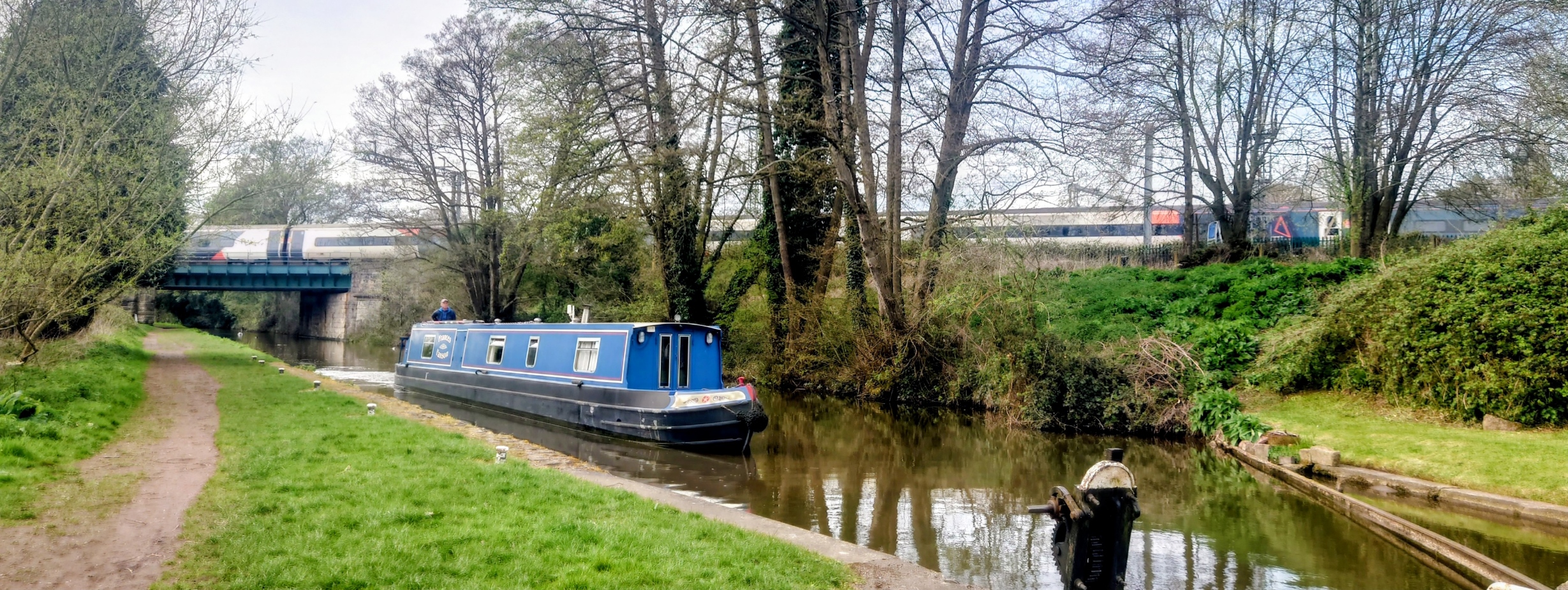 A canal boat navigates as an electric train passes in the background; modern structures don't displace older structures but exists alongside.
